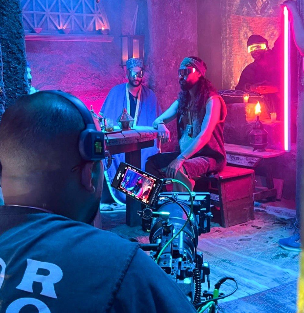 Music video shoot in Morocco — neon-lit traditional Moroccan interior with two performers in shades, cinematographer pulling focus on monitor